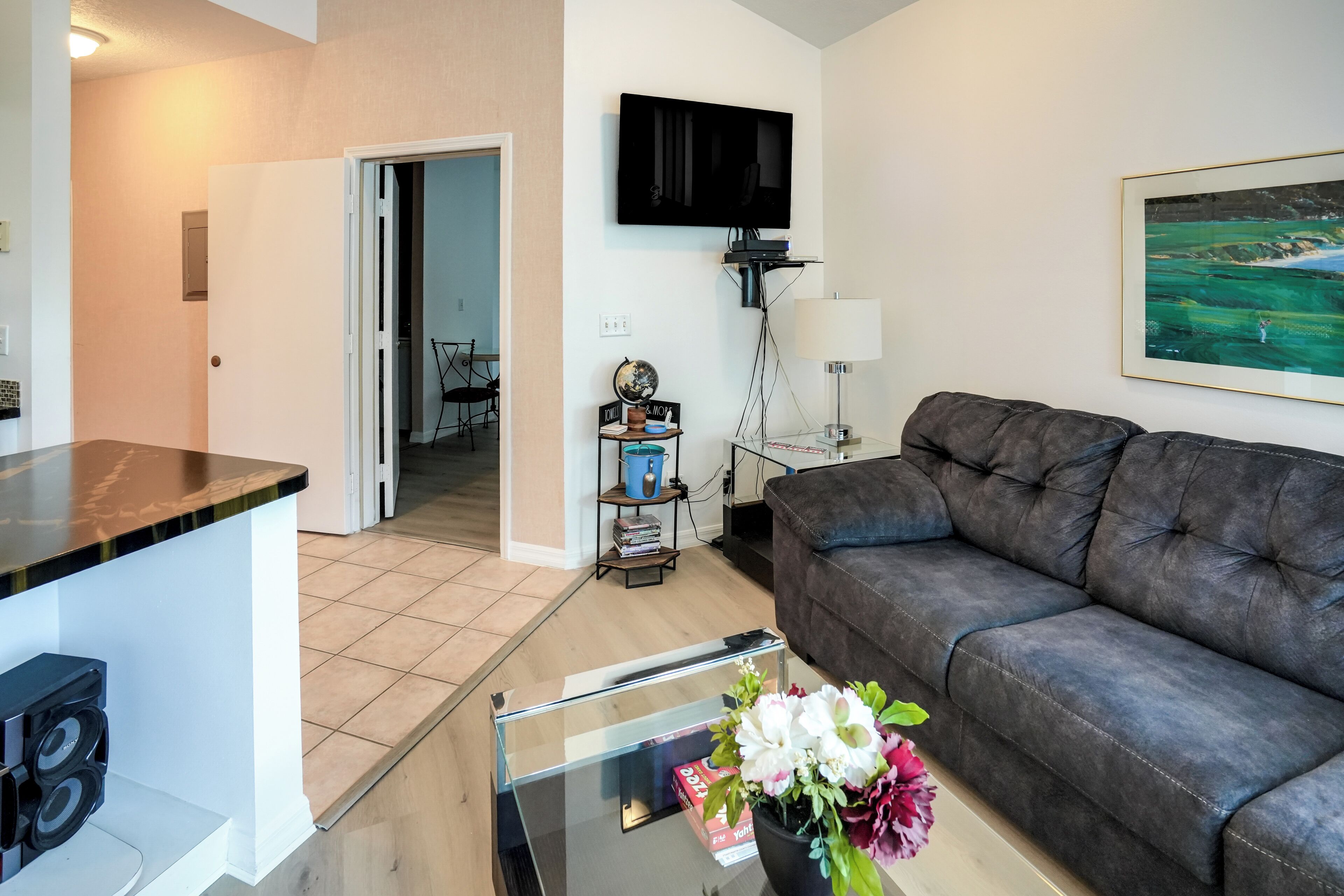 A living room with a dark leather sofa, a glass coffee table with flowers, a flat-screen TV mounted on the wall, and a framed painting.