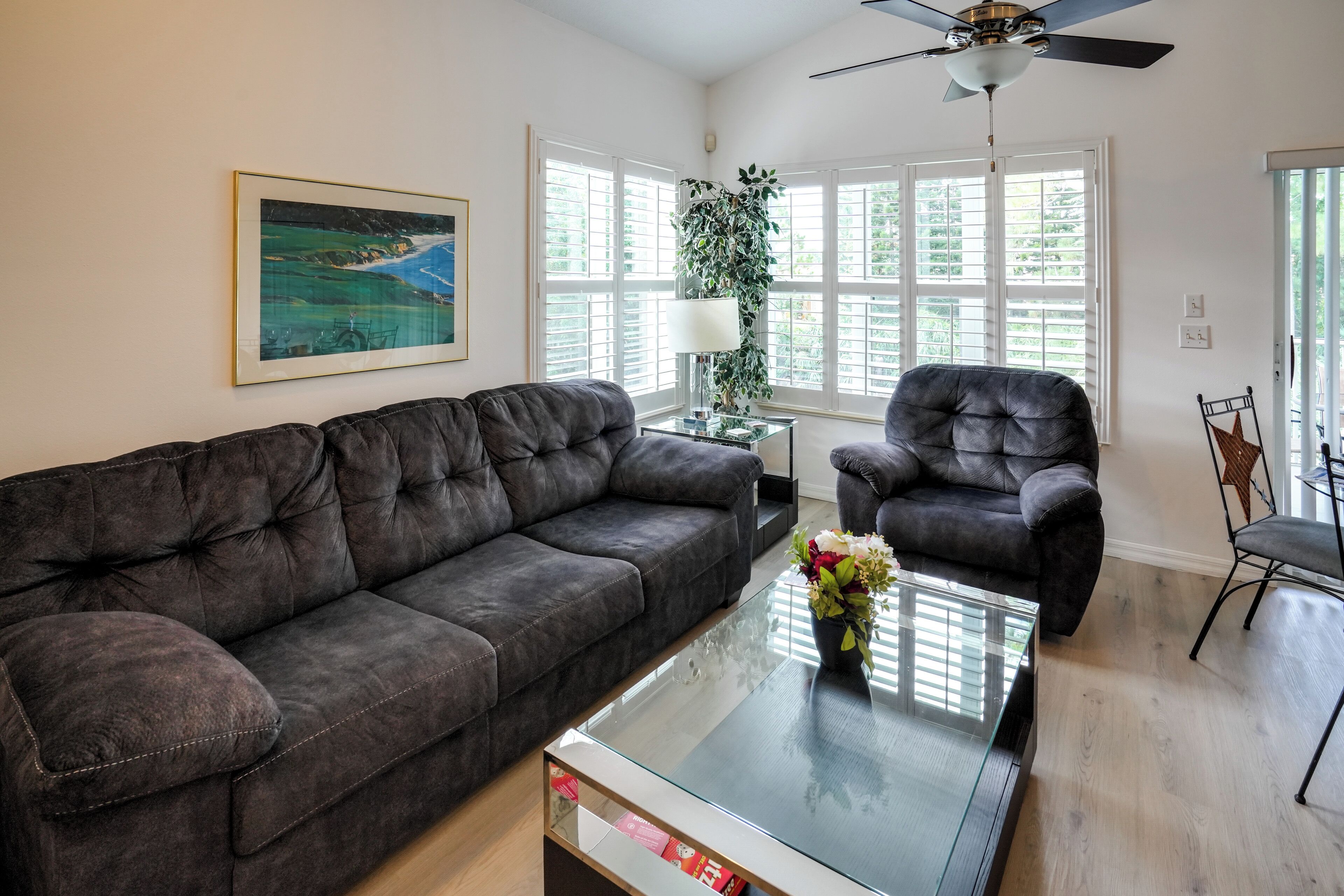 A living room with a dark brown sofa, a glass coffee table with a vase of flowers, a black armchair, and a ceiling fan.