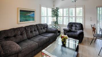 A living room with a dark brown sofa, a glass coffee table with a vase of flowers, a black armchair, and a ceiling fan.