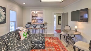 Una sala de estar con un sofá, un reposapiés y una mesita con una lámpara. Se ve una cocina al fondo, con una barra y sillas.