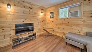 A cozy cabin interior with wooden walls, a television on a wooden stand, a window with a view of trees, and a gray sofa.