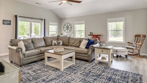 A spacious living room with a large L-shaped sectional sofa, a coffee table, a side table, and a rocking chair. There are three windows providing natural light and a ceiling fan. - Waterfront Townville Home: Hot Tub & Lake Access! (Townville)