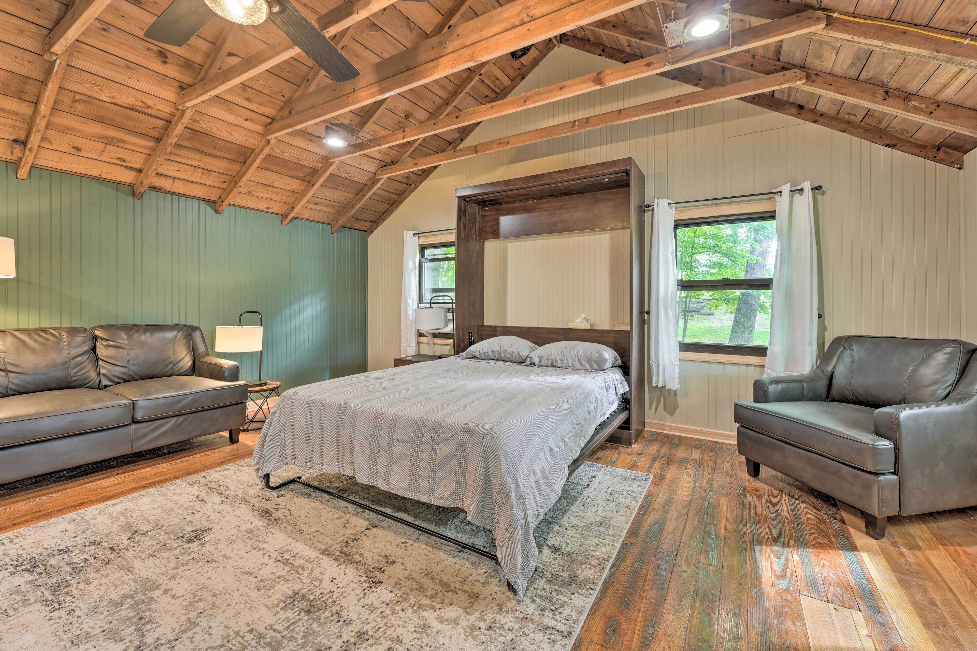A bedroom with a bed, two leather sofas, a wooden ceiling, and a large window.