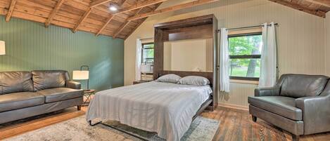 A bedroom with a bed, two leather sofas, a wooden ceiling, and a large window.