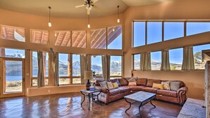A spacious living room with a large leather sectional sofa, a coffee table, and a view of mountains and a lake through large windows.
