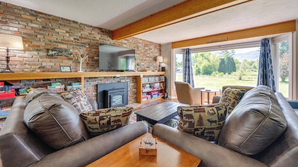 A cozy living room with a brick fireplace, leather sofa, and a large window offering a view of the outdoors. - Peaceful & Private Franconia Home by Cannon! (Franconia)