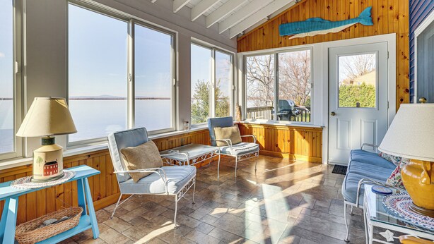 A sunlit room with large windows, a wooden fish wall, and a cozy seating area with two chairs and a table. - Alburgh Lake House: Private Beachfront! (Alburg)