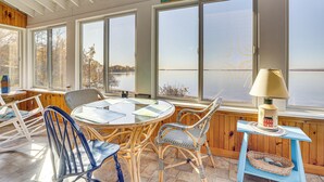 A sunlit room with a large window overlooking a body of water, a table with chairs, and a lamp on a small table. - Alburgh Lake House: Private Beachfront! (Alburg)