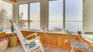 A cozy room with large windows overlooking a lake, wooden furniture, and a view of trees and hills. - Alburgh Lake House: Private Beachfront! (Alburg)
