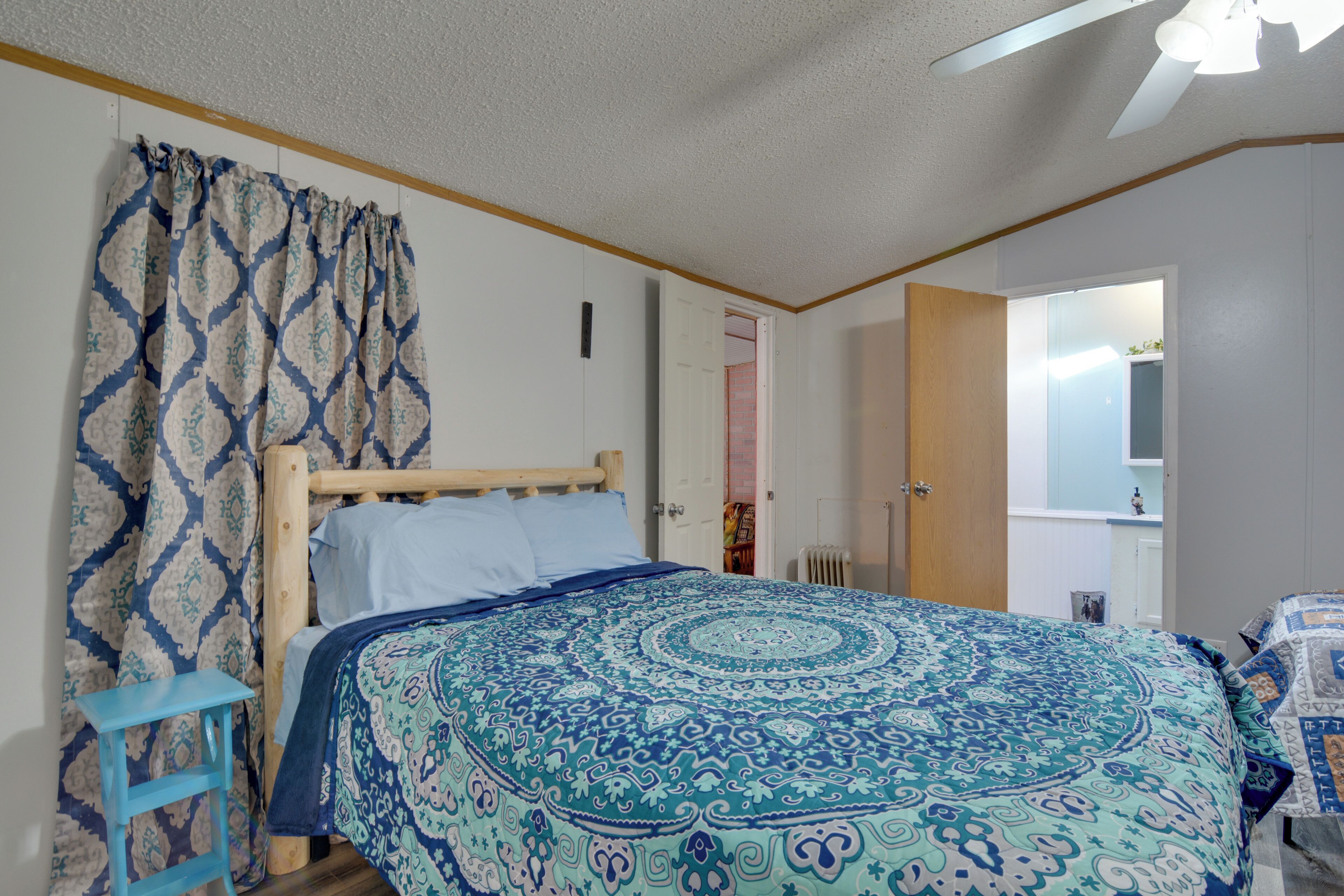 Un dormitorio con cama, cortinas, cabecera de madera, ventilador de techo y una puerta que da a otra habitación.