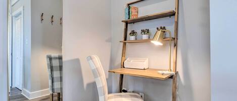 A corner room with a checkered chair, a wooden shelf with books and a lamp, and a mirror reflecting the door and chair.