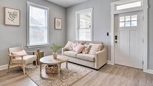 Una sala de estar con un sofá beige, una silla de madera, una mesa redonda y una puerta con un ventanal arriba.