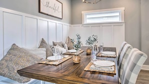 Comedor con mesa de madera dispuesta para cuatro, paredes de paneles blancos, una araña y un cartel enmarcado que dice 'thankful'.