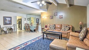 Ein Wohnzimmer mit braunem Ledersofa, einem Couchtisch und einem gemusterten Teppich. Es gibt eine Essecke mit Tisch und Stühlen, und im Hintergrund eine Küche.