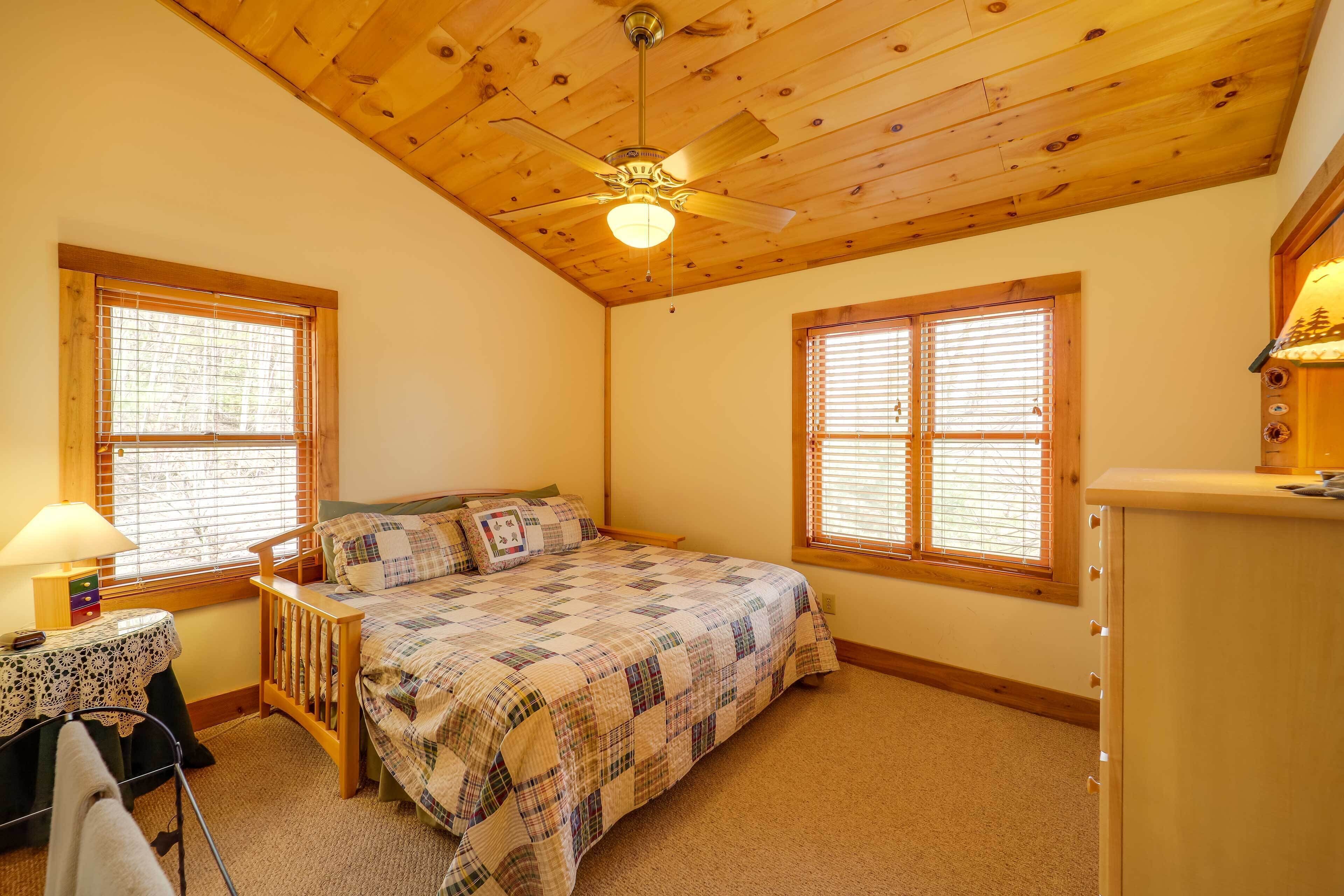 A bedroom with a wooden ceiling, a bed with a quilt, a nightstand, a dresser, and two windows with blinds.