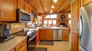 A kitchen with wooden cabinets, stainless steel appliances, and a stone fireplace wall. - Smoky Mountain Vacation Rental w/ Large Deck! (Deep Gap)