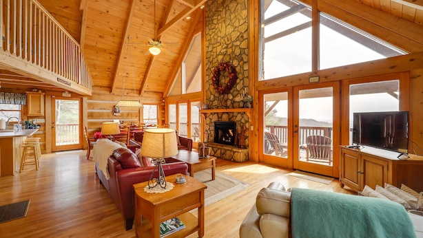 A spacious living area with a stone fireplace, wooden furniture, and large windows. - Smoky Mountain Vacation Rental w/ Large Deck! (Deep Gap)
