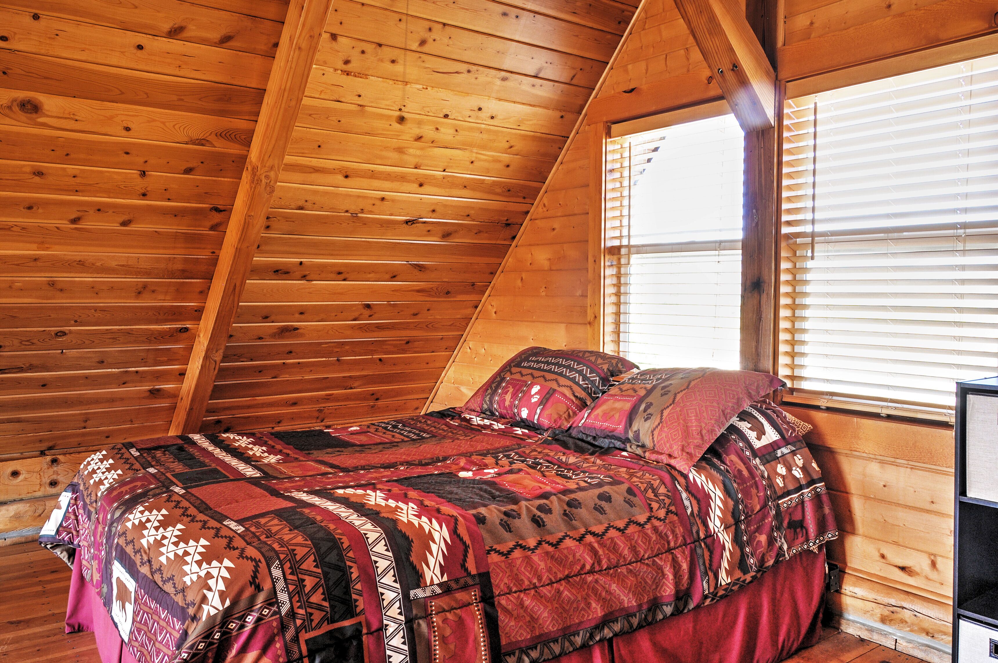 A bedroom with a wooden sloped ceiling, a bed with patterned bedding, and a window with blinds.