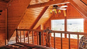 A wooden cabin room with a sloped ceiling, a bed with a patterned blanket, a ceiling fan, and a large window offering a view of the outdoors. - Remote Antimony Log Cabin w/ Green Meadow Views! (Antimony)