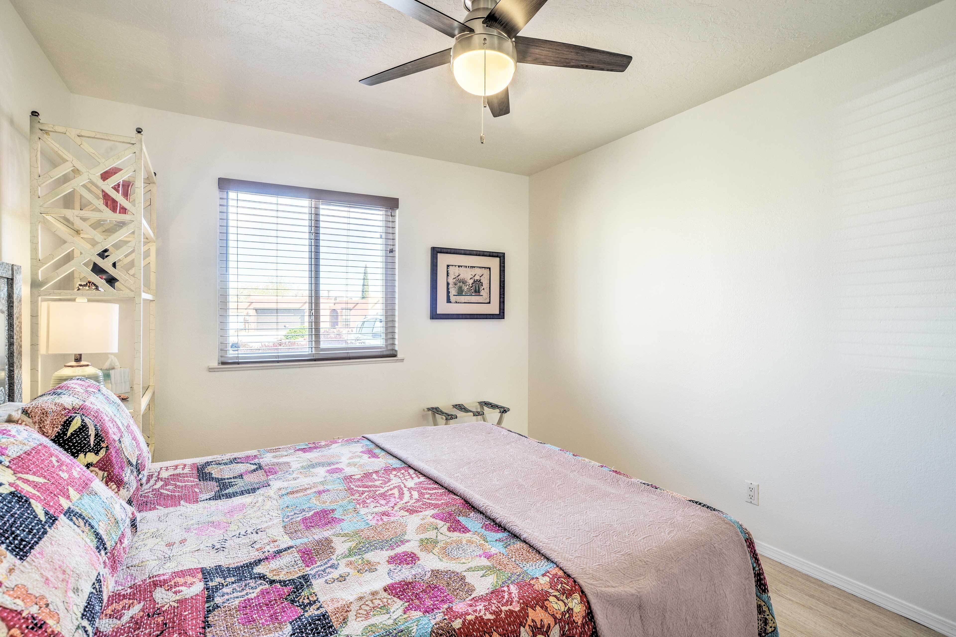A bedroom with a bed, a ceiling fan, a window with blinds, a framed picture on the wall, and a white ladder-like structure against the wall.