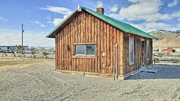 A wooden cabin with a green roof, a window, and a door, set in a gravel area with mountains in the background.