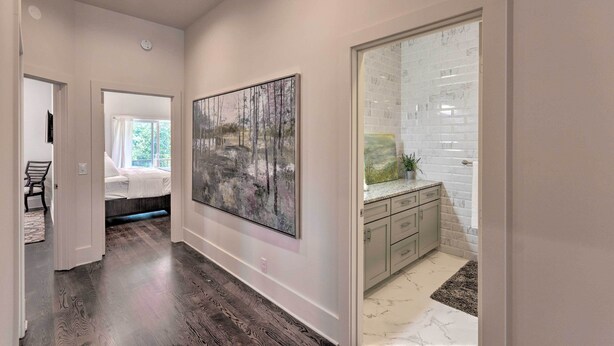 A hallway with a bed, a large abstract painting, a bathroom with a white tiled wall, and a dark wooden floor. - Modern Main St. Apt in Downtown Waynesville! (Waynesville)