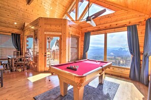 Un intérieur en bois comprenant une table de billard, un coin repas et de grandes fenêtres offrant une vue sur les montagnes.