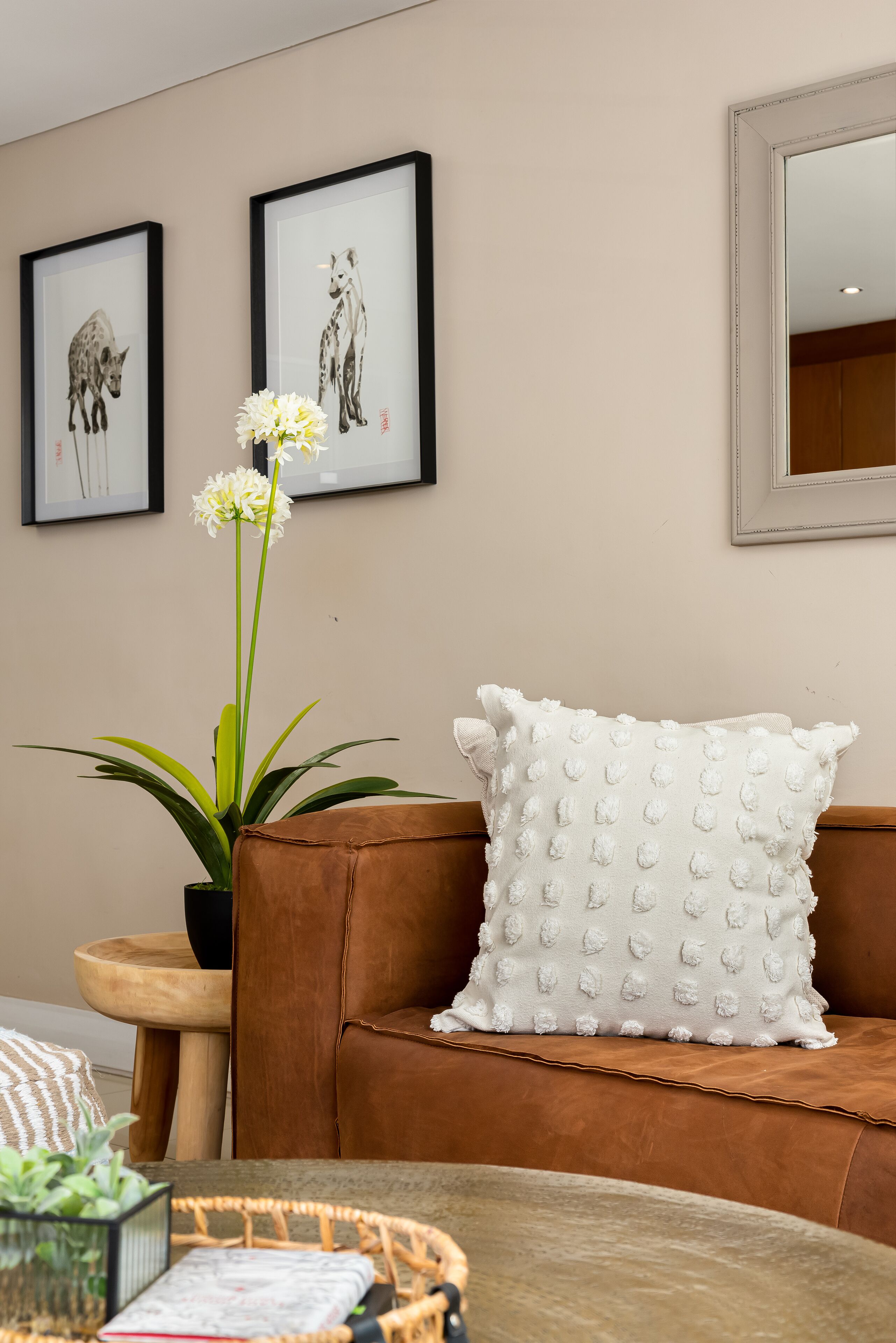 A living room with a brown leather sofa, a white textured pillow, a potted plant with white flowers, and two framed artworks on the wall.