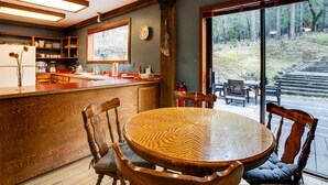A wooden dining table with chairs, a kitchen area with wooden cabinets, and a large window overlooking a forested area. - Peaceful Applegate River Valley Sanctuary! (Grants Pass)