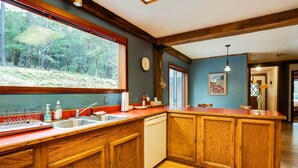 A kitchen with wooden cabinets, a dishwasher, a sink, and a window overlooking a forest. - Peaceful Applegate River Valley Sanctuary! (Grants Pass)