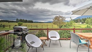 A deck with a grill, two white chairs, a wooden table, and a view of a green landscape with mountains.