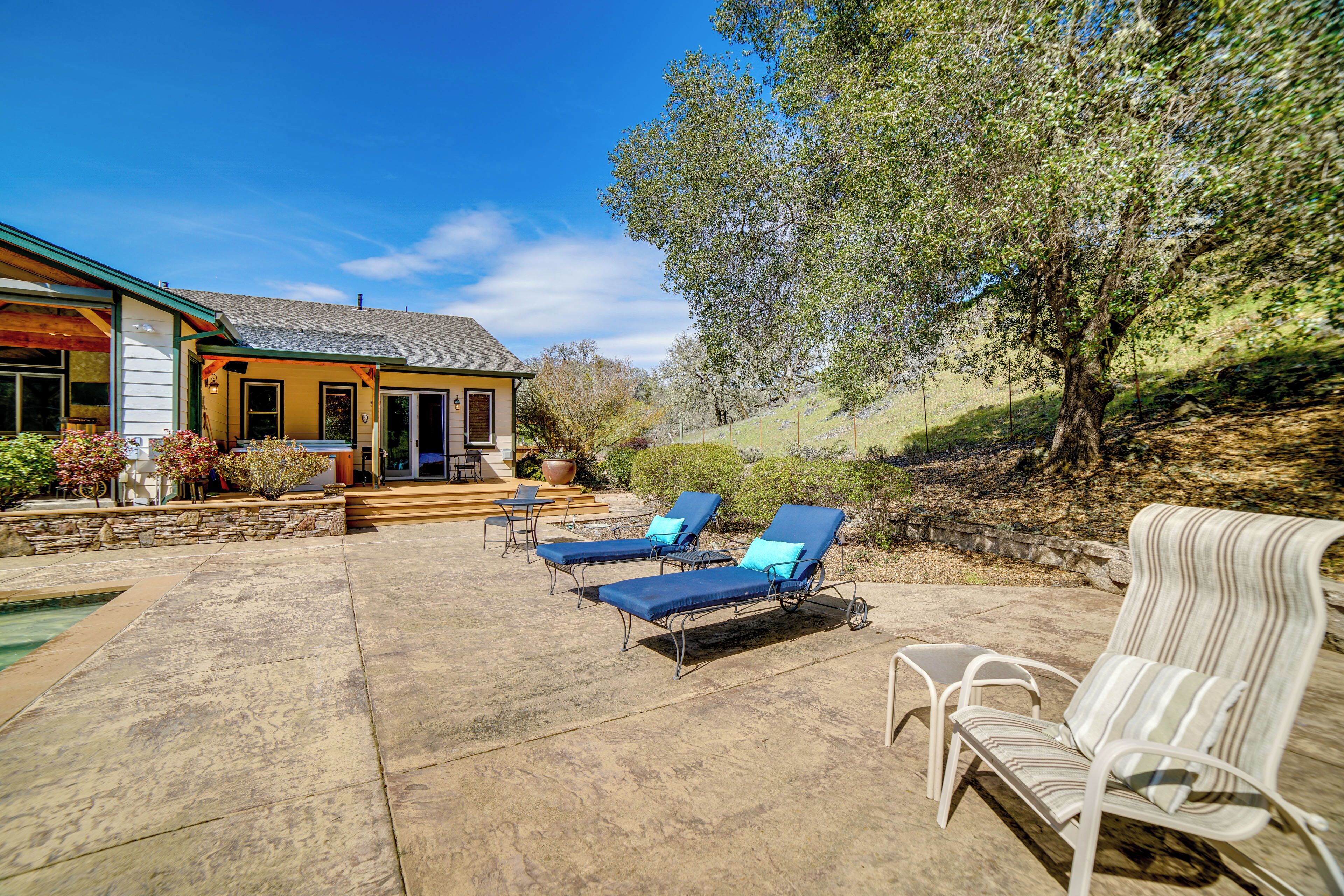A patio with two blue lounge chairs and a white chair, a stone pool, and a house with a yellow exterior and a covered porch.