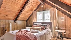 Ein gemütliches Schlafzimmer im Dachgeschoss mit Holzbalkendecke, einem Bett mit rosa Decke, einem Nachttisch mit Lampe und einem Fenster mit Vorhängen.