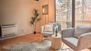 Ein gemütliches Zimmer mit großem Fenster, einer Topfpflanze, einem grauen Sessel, einem kleinen runden Tisch und einem weißen Heizkörper.