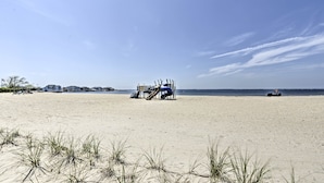 Una playa con una sombrilla azul y una silla, una zona arenosa con césped, y un cielo despejado.