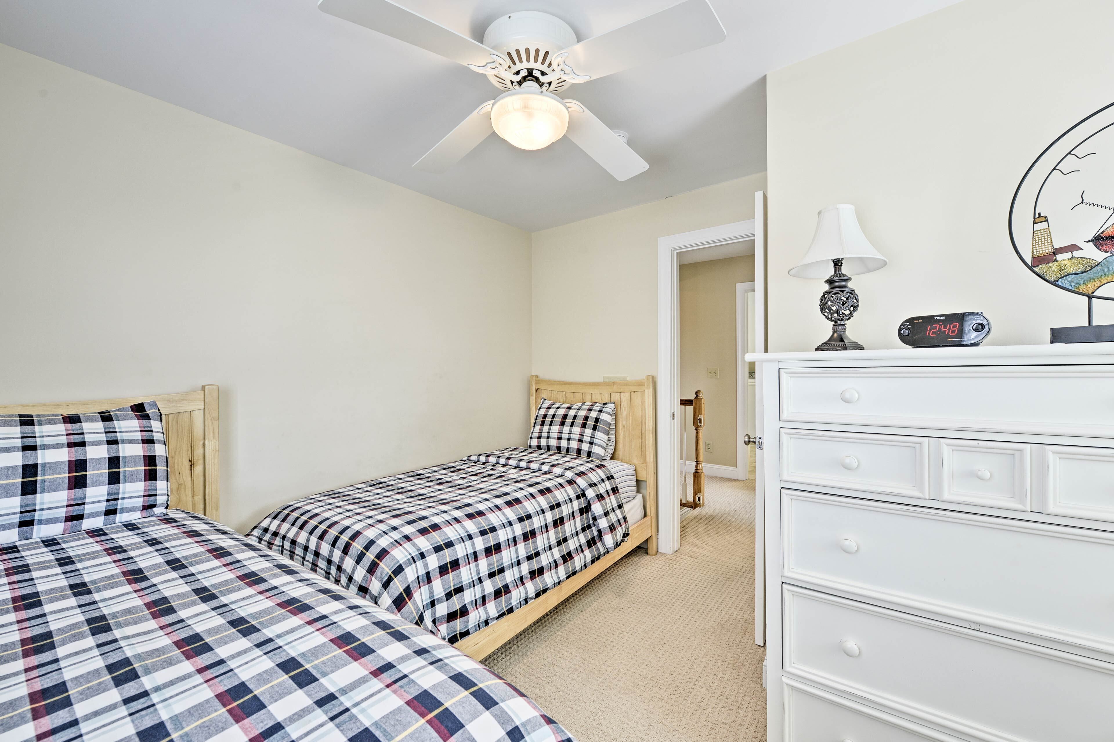 Quarto com duas camas, um ventilador de teto, cômoda branca, luminária e relógio.