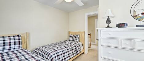 Quarto com duas camas, um ventilador de teto, cômoda branca, luminária e relógio.
