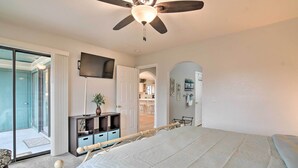 A bedroom with a bed, a ceiling fan, a TV, a sliding glass door, a small desk, and a plant.