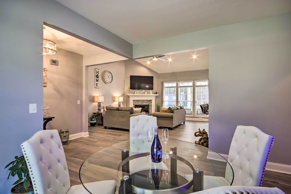 A modern living room with a glass coffee table, white upholstered chairs, a fireplace, and a television mounted above it.