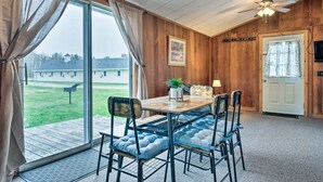Zona pranzo con tavolo e sedie in legno, ventilatore a soffitto e vista su un prato attraverso ampie finestre.