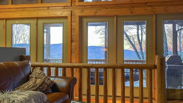 Interior de una cabaña con piso de madera, un sofá de cuero y ventanas amplias que ofrecen vistas de un paisaje nevado.