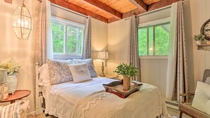 A cozy bedroom with a wooden ceiling, a bed with white bedding, a bedside table with a lamp, and a window with curtains. - Welcoming Poconos Retreat w/ Deck & Pool Access (Tobyhanna)