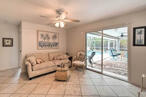 A living room with a beige sofa, a chair, a glass coffee table, and a ceiling fan. There is a large window with a view of a pool and outdoor furniture.