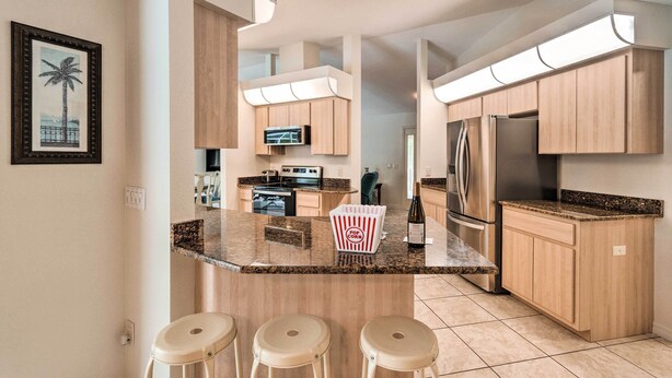 A modern kitchen with wooden cabinets, a granite countertop, and a built-in oven. There are three stools at the island and a framed picture on the wall. - Englewood Escape w/ Pool ~ 6 Mi to Beaches! (Englewood)