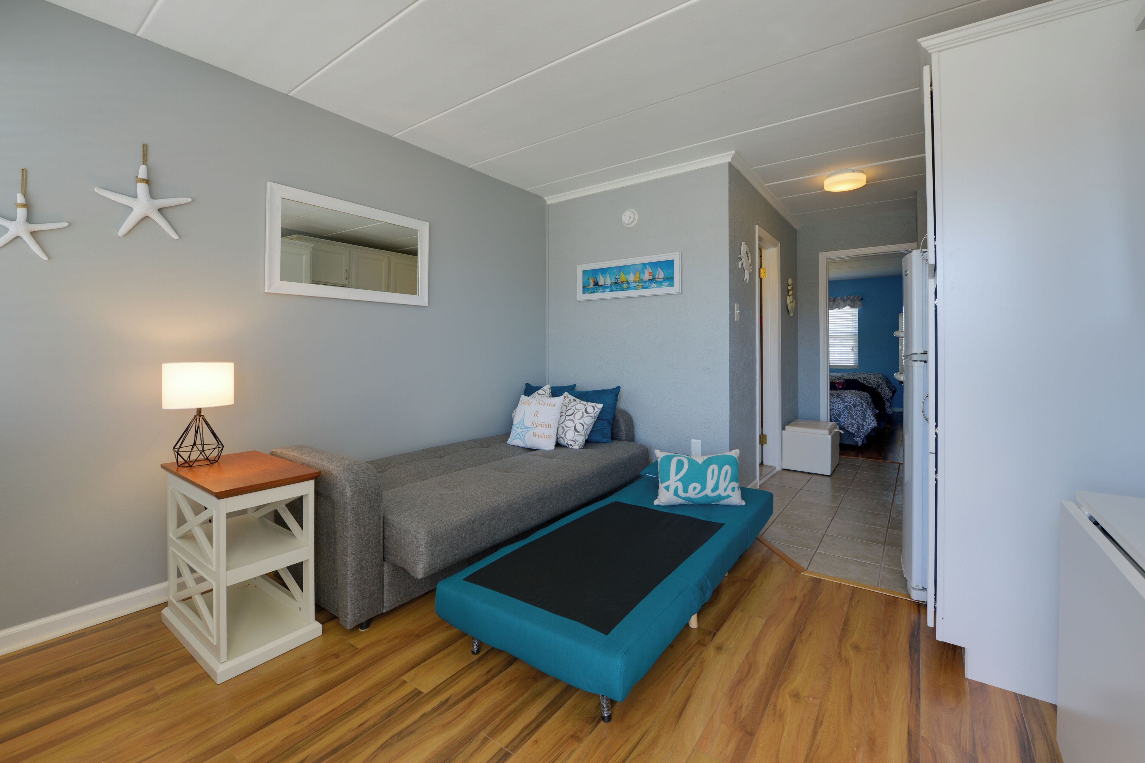 A bedroom with a bed, a small table with a lamp, a bench, and a door leading to another room.