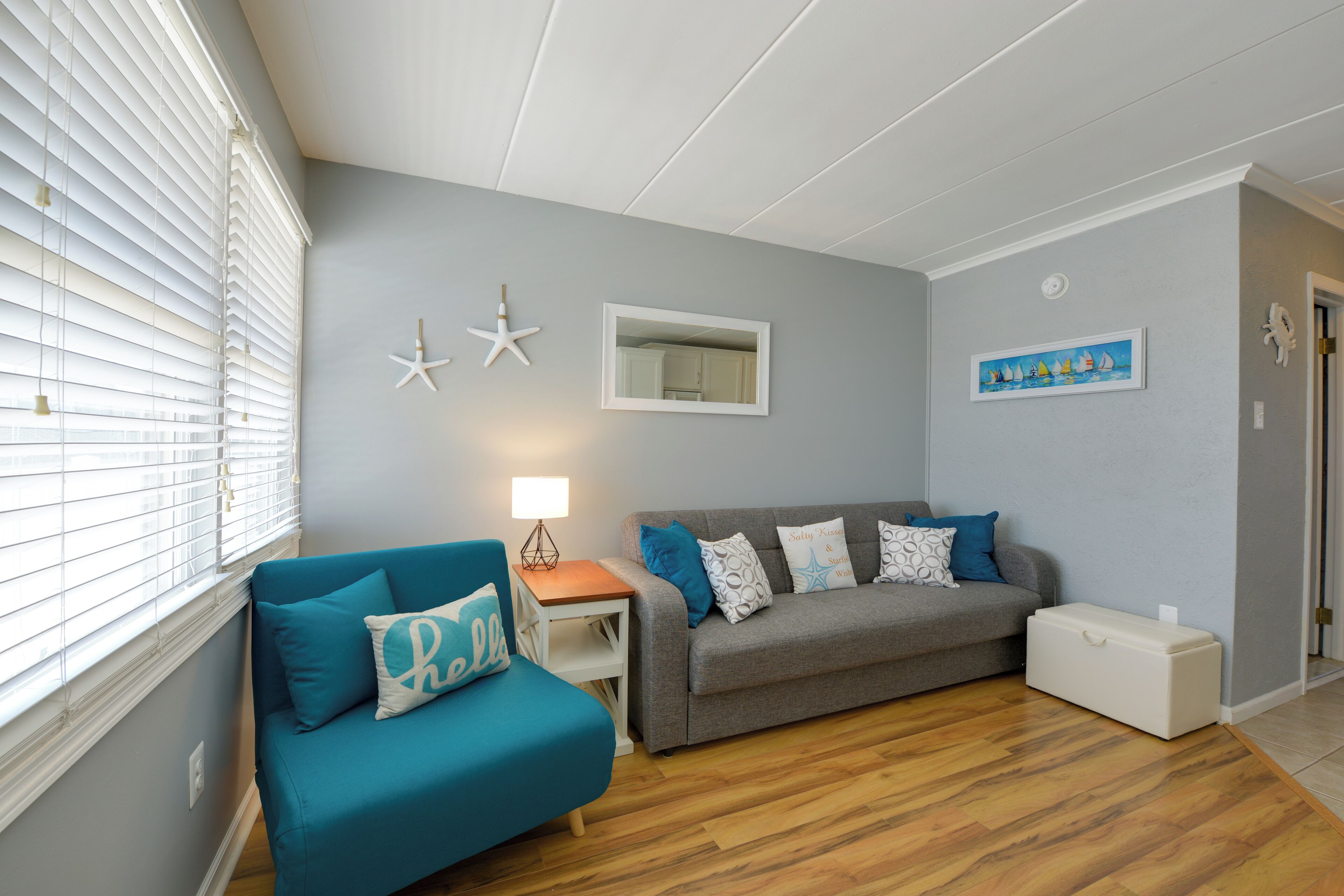 A living room with a teal sofa, a grey sofa, a wooden floor, a small table with a lamp, and a white storage box.
