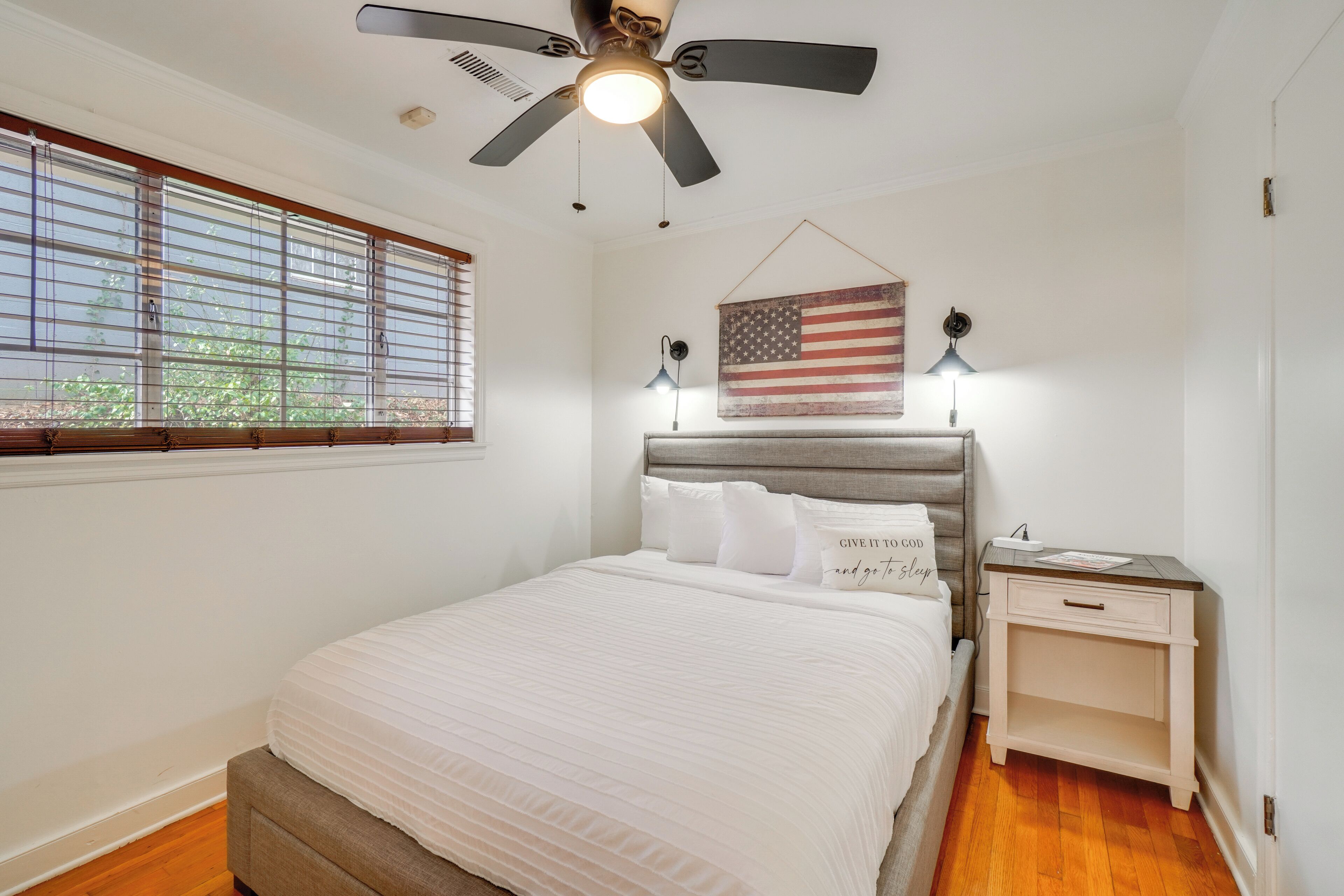 A bedroom with a bed, a nightstand, a ceiling fan, a window with blinds, and an American flag decoration.