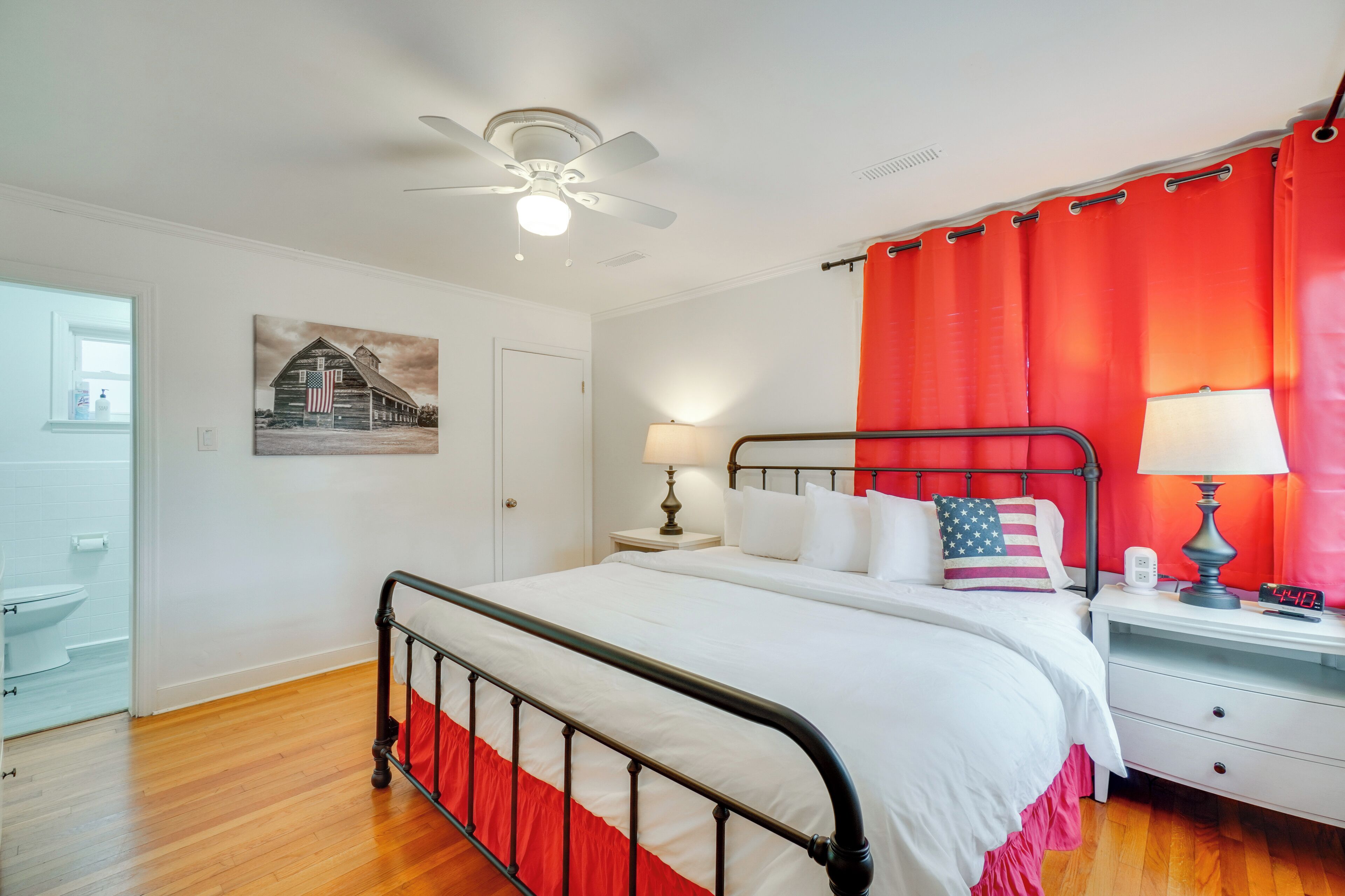 Une chambre à coucher comprenant un lit, des tables de chevet, un ventilateur de plafond, une fenêtre avec des rideaux, un tableau accroché au mur et une salle de bain visible à travers une porte ouverte.