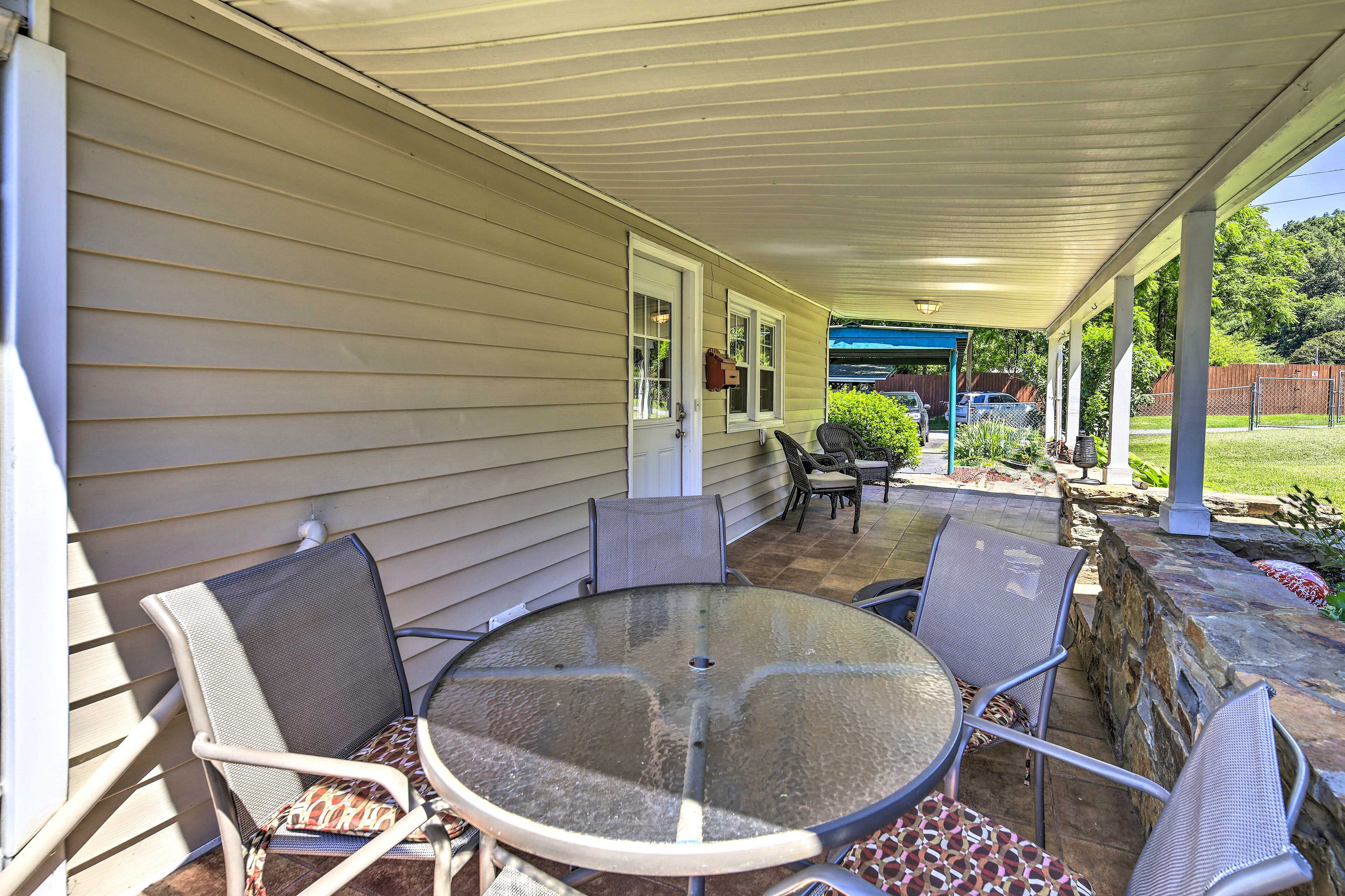 An outdoor patio with a round glass table and four chairs, a covered area with a roof, and a stone wall.