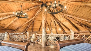 Ein rustikaler Holzinnenraum mit hoher Decke, freiliegenden Balken und einer zentralen Holzsäule. Es gibt zwei Holzbetten mit blau gestreifter Bettwäsche.